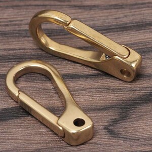 May include: Two brass-colored carabiner clips are displayed on a wooden surface. The clips have a spring-loaded gate and a small hole at the base. The carabiners are designed for securing items and are commonly used in various applications.