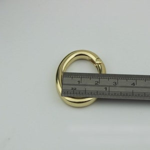 May include: A gold-colored metal oval ring with a swivel clasp, measured against a ruler. The ring is approximately 2 inches in diameter.
