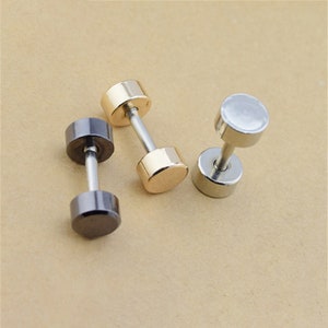 May include: Three dumbbell-shaped stud earrings in the image. One is black, one is gold, and one is silver. The earrings have a cylindrical shape with a post in the center. The earrings are on a light brown surface.