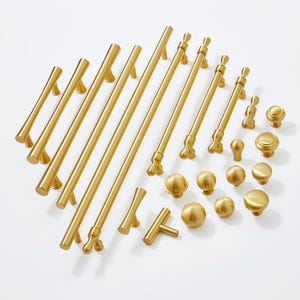 May include: A collection of gold-toned cabinet hardware, including knobs and pulls in various styles and sizes. The hardware is arranged on a white surface.