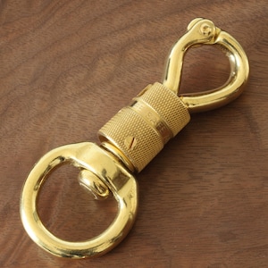 May include: A gold-colored swivel snap hook with a textured center section and two rounded loops. The words "BE SAFE" are engraved on the center section. The hook is designed to rotate and connect items.