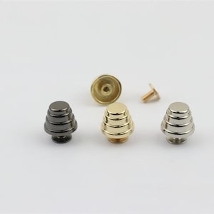 May include: Four metal pyramid-shaped studs in black, gold, and silver. The studs have a layered design and are used for attaching to leather goods.