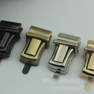 May include: Four metal purse clasps in different finishes: black, antique brass, silver, and gold. Each clasp has a rectangular body with a curved design and a small tab. These clasps are likely used for bags or other accessories.