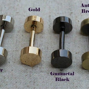 May include: Four dumbbell-shaped stud earrings in silver, gold, gunmetal black, and antique bronze. Each earring has a cylindrical post connecting two flat, round discs. The text labels each color: Silver, Gold, Gunmetal Black, and Antique Bronze.