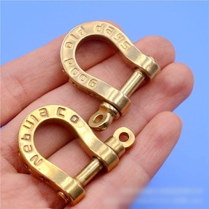 May include: Two brass-colored D-shackles with a polished finish. Each shackle has a curved top with engraved text and a removable pin. The text on one shackle reads "Old Poog 2006" and the other reads "Nequila Co".