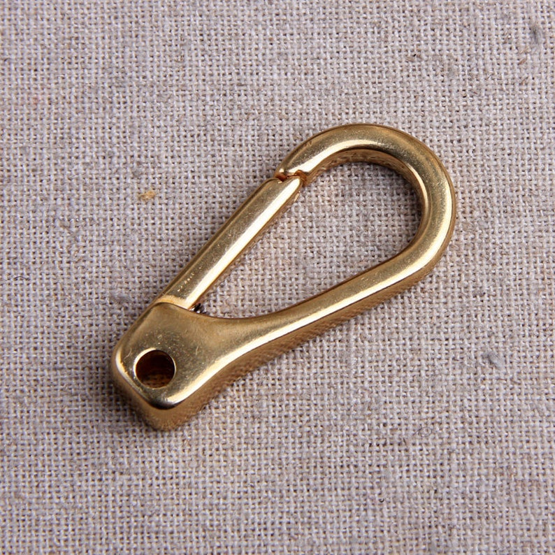 May include: A gold-colored spring snap hook, also known as a carabiner, is displayed against a neutral fabric background. The hook has a rounded shape with a spring-loaded gate and a small hole at the base. This hardware item is used for various applications.