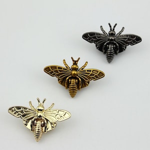 May include: Three bee-shaped metal embellishments in gold, silver, and black. The bees have detailed wings and bodies.