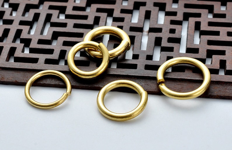 May include: Five gold-colored metal jump rings of varying sizes. The rings are smooth and shiny.