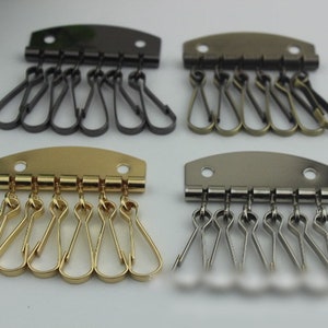 May include: Four key holder hardware pieces in various metallic finishes: black, bronze, gold, and silver. Each piece has a curved top with two holes and multiple key rings attached. Ideal for organizing keys.