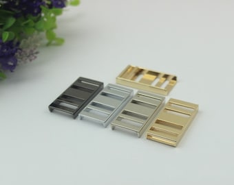 Può includere: Una collezione di fibbie rettangolari in metallo in varie finiture: nero, argento e oro. Ogni fibbia ha un design a fessura con tre aperture orizzontali. Le fibbie sono probabilmente destinate all'uso in borse, cinture o altri accessori.