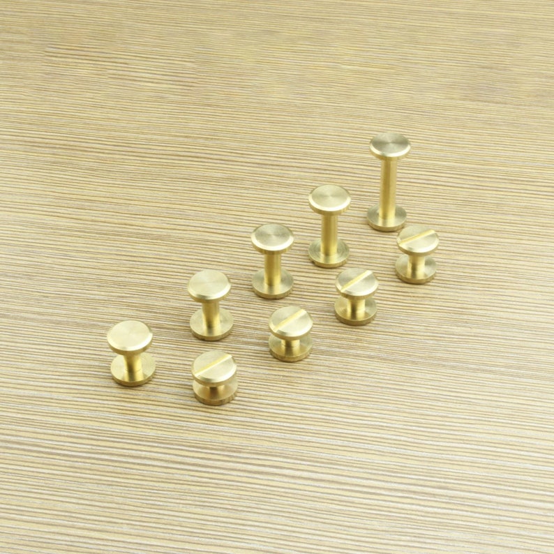 May include: A collection of ten brass screw-in studs, arranged in a pyramid shape on a wooden surface. The studs are all the same size and have a round head with a small screw on the bottom.