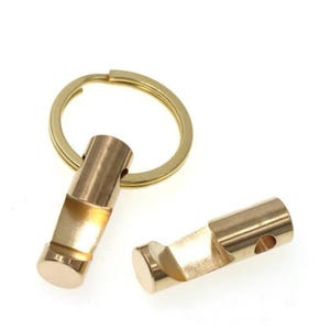 May include: Two brass bottle opener keychains. One keychain has a gold ring attached. The bottle openers are cylindrical with a slot for opening bottles. The keychains are on a white background.