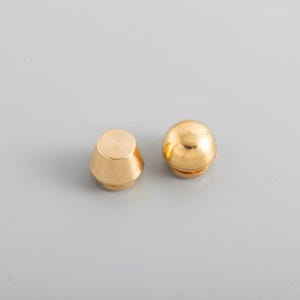 以下が含まれることがあります： グレーの背景に、2つの小さく光沢のある金色の金属製のオブジェクトがあります。1つは平らな上面を持つ丸みを帯びた円錐形で、もう1つは球体です。これらのオブジェクトは、家具や工芸品用のハードウェアコンポーネントのようです。