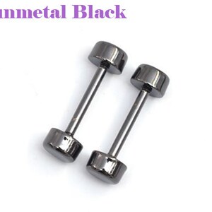 May include: Two gunmetal black dumbbell-shaped earrings. Each earring has a cylindrical post connecting two rounded, flat-topped cylinders. The text "Gunmetal Black" is in the upper left corner. The earrings are set against a white background.