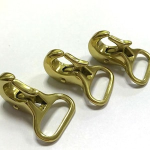 May include: Three brass-colored swivel snap hooks. Each hook has a spring-loaded clasp and a D-ring. The hooks are arranged on a white surface, with the light reflecting off the polished metal.