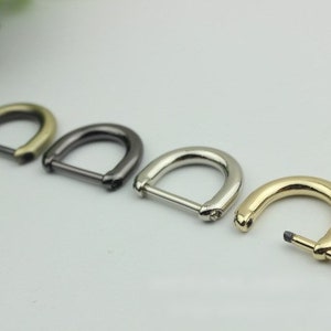 May include: Four metal D-rings in various finishes: antique brass, dark gunmetal, silver, and gold. The D-rings are arranged in a row, with one partially disassembled, showing the pin. These hardware pieces are likely for bags or other accessories.