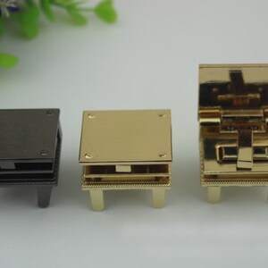 May include: Three metal clasps for bags or purses. The clasps are in different finishes: black, gold, and gold with a hinged lid. The clasps have a square shape and four legs.