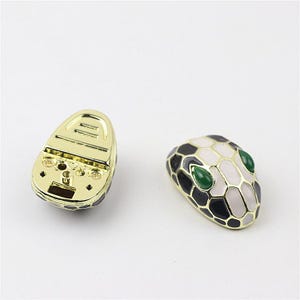 May include: A pair of gold-tone snake head cufflinks. One cufflink is a detailed snake head with black and white scales and green eyes. The other cufflink is the back of the snake head, showing the gold-tone metal.