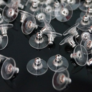 May include: A close-up of a group of clear plastic earring backs with silver metal posts. The backs are designed to be used with stud earrings.