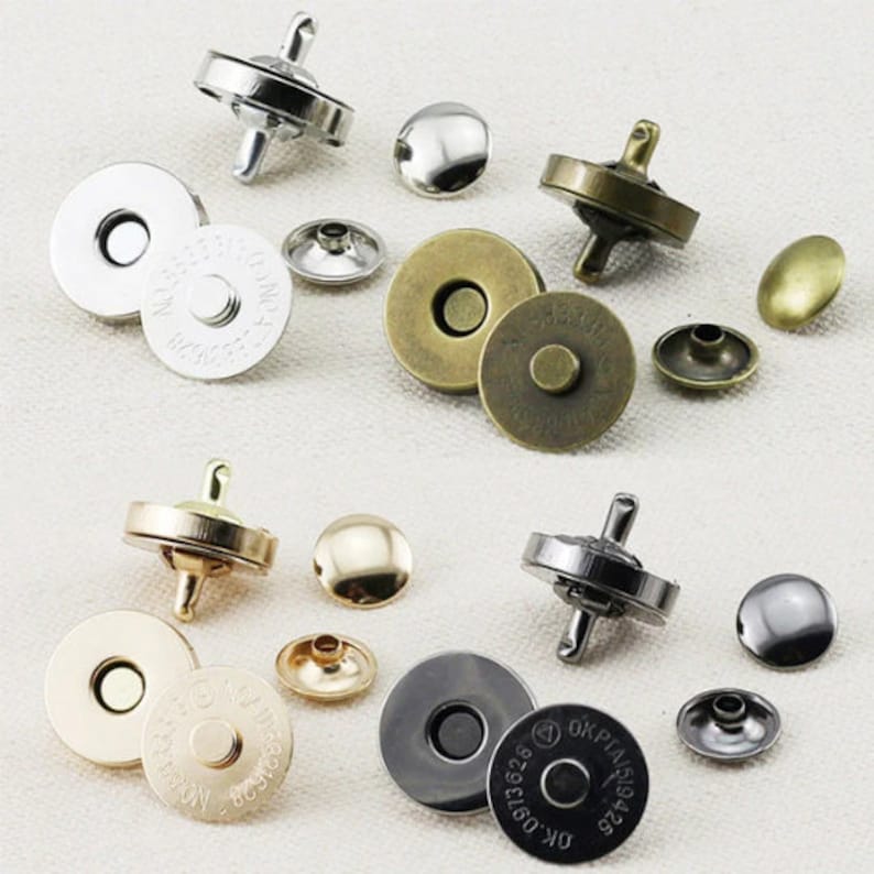 May include: A selection of magnetic snap closures in silver, gold, and bronze finishes. The closures are round and flat, with a central hole for sewing. Some have a loop for attaching to a bag or garment. The image shows the different parts of the snap.