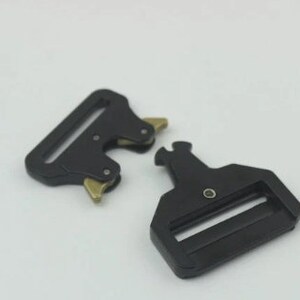 May include: Two black metal belt buckle clips with a gold finish. The clips are designed to be used with webbing straps.