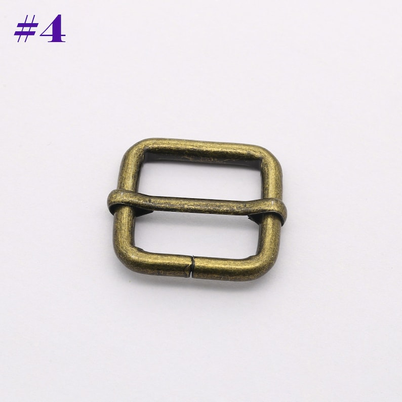 May include: A gold-toned metal buckle with a square shape and a center bar. The buckle is designed for use with straps or belts.