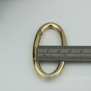 May include: Gold-toned oval-shaped metal clasp with a spring-loaded mechanism. The clasp is measured with a ruler, showing it is approximately 2.5 inches long.