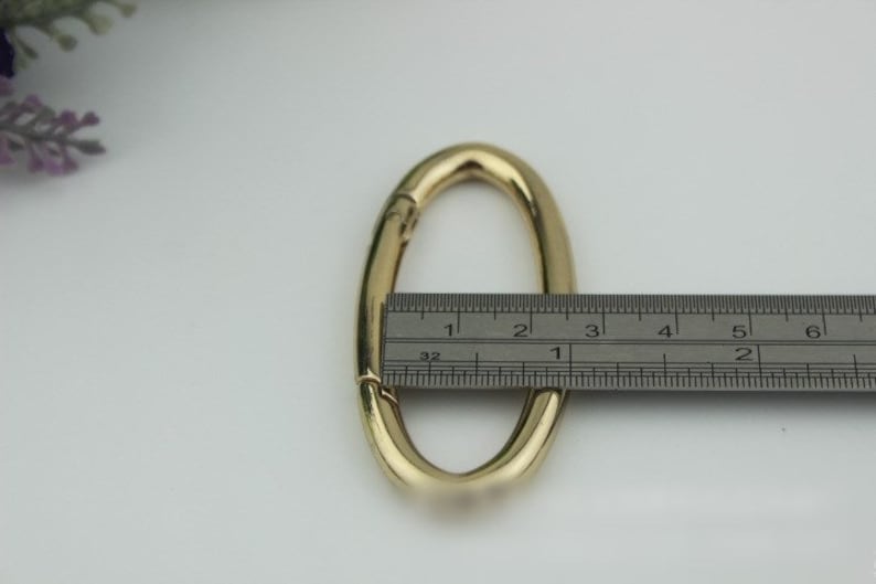 May include: Gold-toned oval-shaped metal clasp with a spring-loaded mechanism. The clasp is measured with a ruler, showing it is approximately 2.5 inches long.