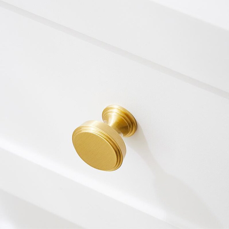 Op de afbeelding: Een ronde, goudkleurige kastknop met een licht verhoogde rand. De knop heeft een eenvoudig, elegant ontwerp.