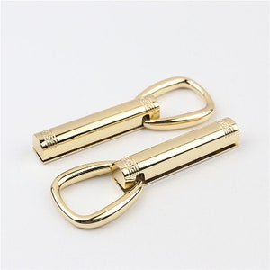 May include: Two gold-toned metal clasps with a ring on one end. The clasps have a decorative pattern.