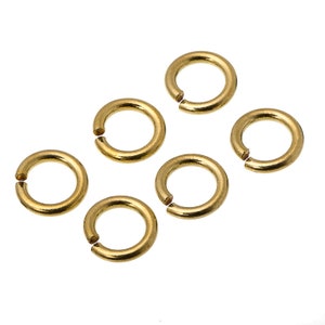 May include: Six gold jump rings, also known as split rings, are arranged on a white background. These rings are commonly used in jewelry making and crafting.