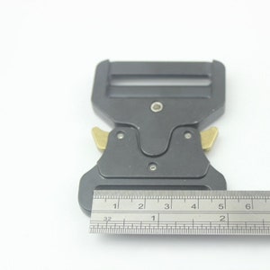 May include: A black metal buckle with two gold-colored release levers. The buckle is 2 inches long and is measured with a ruler.