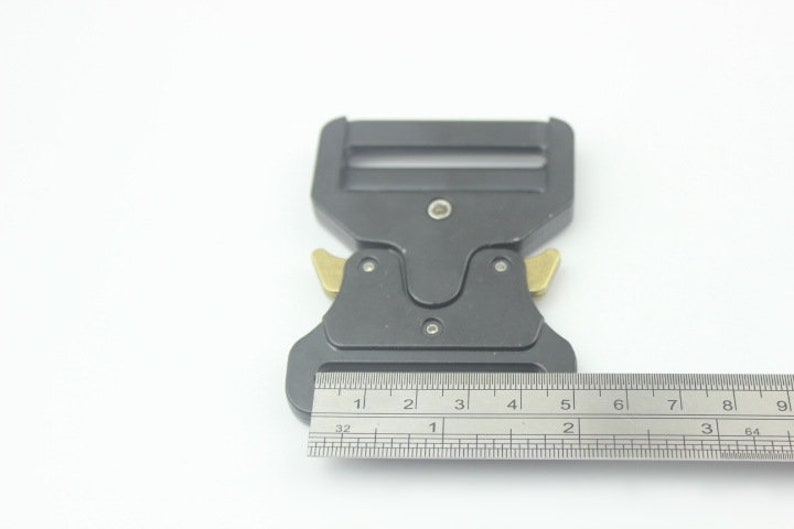 May include: A black metal buckle with two gold-colored release levers. The buckle is 2 inches long and is measured with a ruler.