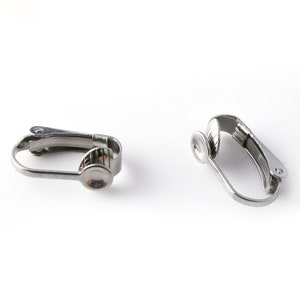 Könnte beinhalten: Zwei silberfarbene Ohrclips. Die Clips sind so konzipiert, dass sie Ohrringe für Menschen mit empfindlichen Ohren an Ort und Stelle halten.