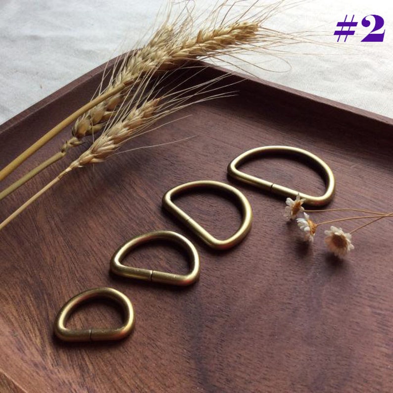 May include: Four brass D-rings of different sizes on a wooden surface. The rings are arranged in order of size, with the largest ring at the top and the smallest ring at the bottom.