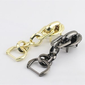 May include: Two metal chain clasps, one gold and one black, with a D-ring on each end. The clasps are designed for attaching to bags or other items.