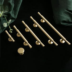 Op de afbeelding: Een set van zeven gouden kastgrepen met een cilindrische vorm en afgeronde uiteinden. De grepen zijn in een rij op een zwart oppervlak gerangschikt.