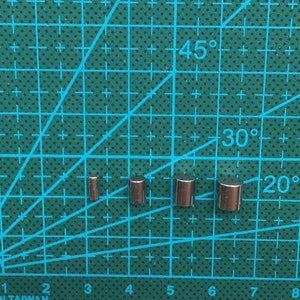 May include: Four silver cylindrical magnets of varying sizes on a green cutting mat with a grid pattern. The mat has markings for angles in degrees.