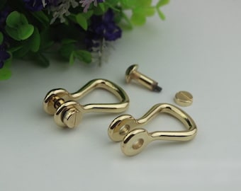 May include: Two gold-colored U-shaped metal clasps with screws, along with two additional screws. The clasps have a shiny finish and are designed for attaching straps or other hardware. The background includes green and purple foliage.