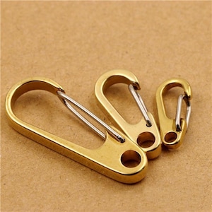 May include: Three gold-toned metal carabiner clips of varying sizes, each featuring a smooth, polished finish and a silver-colored spring-loaded gate mechanism. The clips have an elongated, rounded shape with a circular hole at the base.