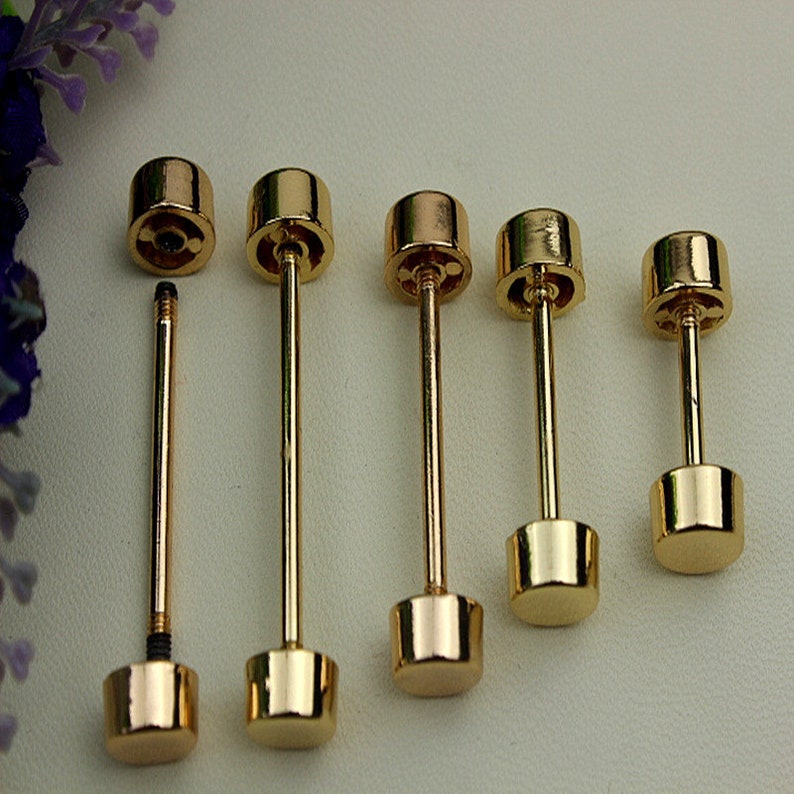 May include: A collection of five gold-colored tie tacks. Each tack features a cylindrical top and a square-shaped base, connected by a slender, straight post. One tack is disassembled, showing the threaded post and the separate top.