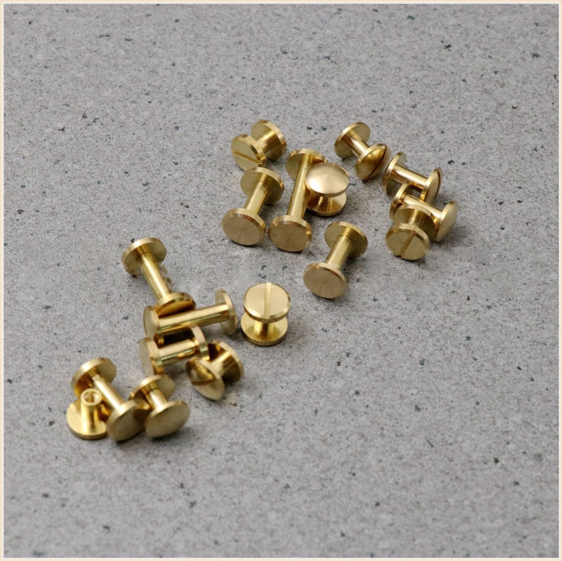 May include: A group of 15 brass screw-on studs, also known as rivets, scattered on a grey surface. The studs are made of a shiny, gold-coloured metal.