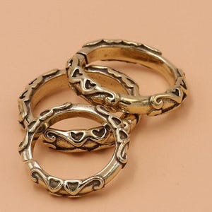 May include: Three antique brass rings with an ornate heart and scroll design. The rings are stacked on a peach-colored surface, showcasing their intricate details and vintage aesthetic. The rings are made of brass.