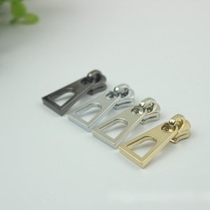 May include: Four zipper pulls in different finishes: black, silver, nickel, and gold. Each pull is a rectangular shape with a small opening in the center.