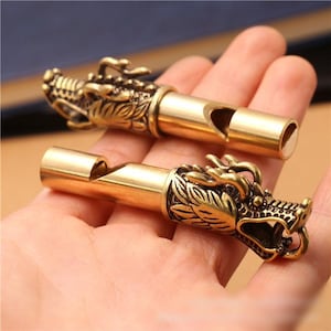May include: Two cylindrical brass whistles, each featuring a detailed, three-dimensional dragon head sculpture at one end. The metal has a polished, golden-yellow finish with darkened recessed areas that highlight the intricate scales and facial features.