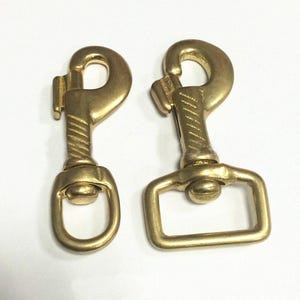 May include: Two gold-toned metal swivel snap hooks with a polished finish. The left hook features a round swivel eye, while the right hook has a rectangular swivel eye. Both fasteners include a spring-loaded gate mechanism and a textured neck.