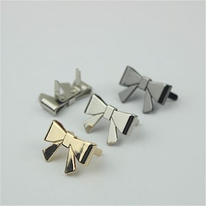 Peut inclure: Trois embellissements en métal en forme de nœud, en or, argent et gris foncé. Les nœuds ont un design plat, bidimensionnel et sont destinés à être fixés à du tissu ou à d'autres matériaux.