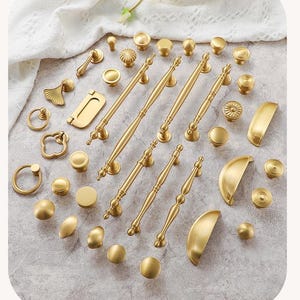 May include: A collection of gold-toned cabinet hardware, including knobs, pulls, and handles. The hardware features various designs, from simple round knobs to ornate handles with decorative detailing. The items are arranged on a light-colored surface.