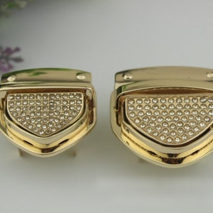 May include: Two gold-toned metal clasps with a diamond-shaped design. Each clasp has a decorative, raised, diamond-shaped section with small, clear rhinestones.