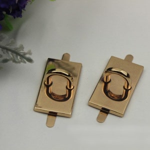 May include: Two gold-toned metal rectangular handbag hardware pieces with a ring in the center. Each piece has a small metal tab on the top and bottom.